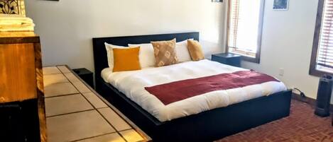 A bedroom with a bed, a bedside table, a bench, and a window with blinds.