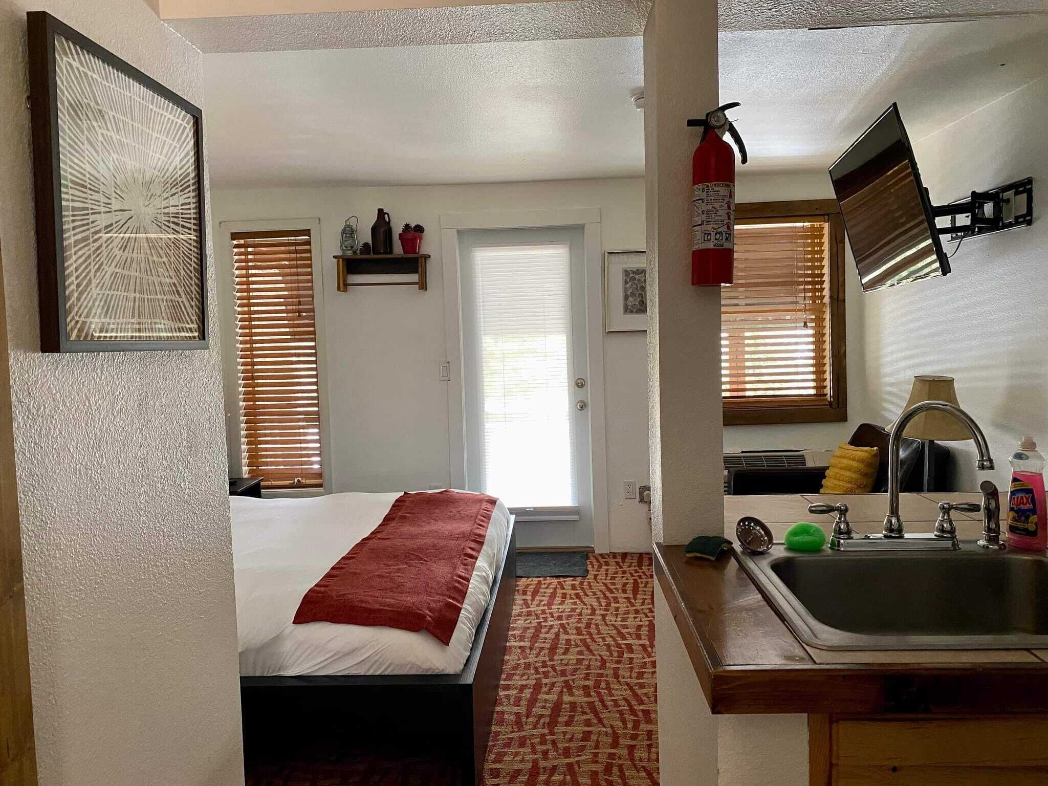 A compact hotel room with a bed, a small kitchenette, a bathroom, and a door leading to another room.
