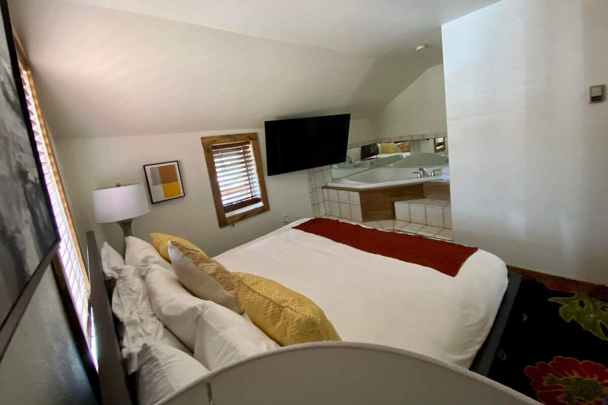 Una camera d'albergo con un letto grande, un comodino, una lampada, una finestra con persiane, un televisore montato a parete e un bagno visibile attraverso una porta aperta.