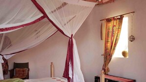 A bedroom with a bed under a mosquito net, a bedside table, a suitcase, and a window with curtains. - Bugesera Lodge (Ntarama)