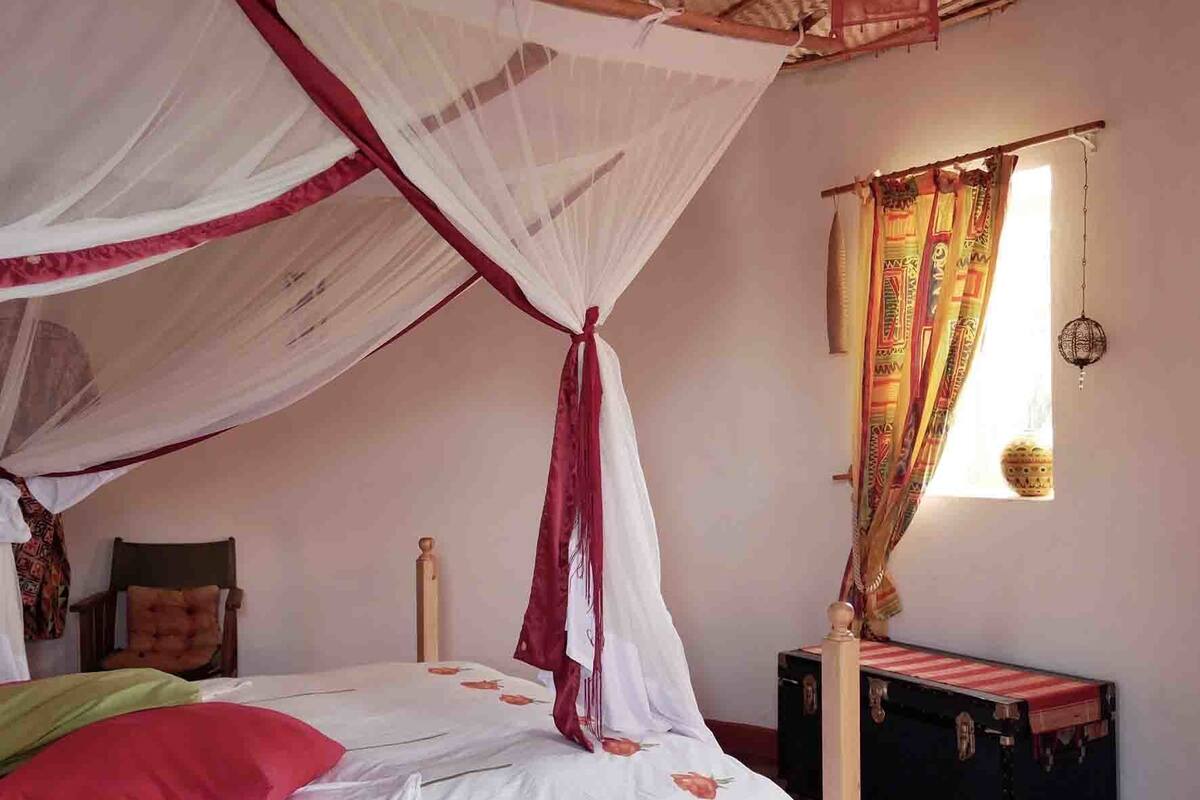 A bedroom with a bed under a mosquito net, a bedside table, a suitcase, and a window with curtains.