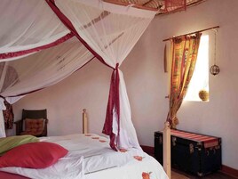 Quarto com cama sob um mosquiteiro, mesa de cabeceira, mala e janela com cortinas.