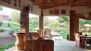 A covered outdoor seating area with brick pillars, a thatched roof, and a view of a garden with potted plants and a lawn. - Bugesera Lodge (Ntarama)