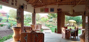 Uma área de estar coberta ao ar livre, com pilares de tijolo, telhado de palha e vista para um jardim com plantas em vasos e gramado.
