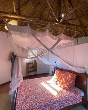 A room with a bed under a mosquito net, a wooden headboard, and a shelf with books. - Bugesera Lodge (Ntarama)
