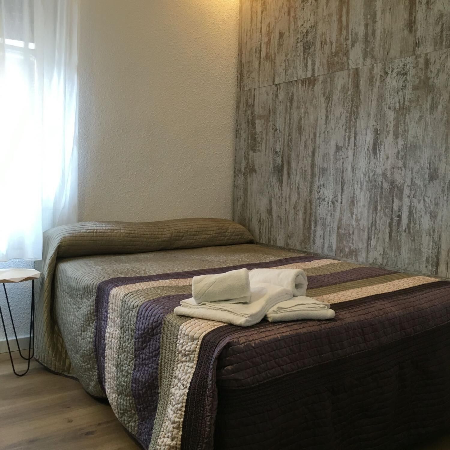 A neatly made bed with a striped blanket and folded towels, a small bedside table, and a wooden textured wall.