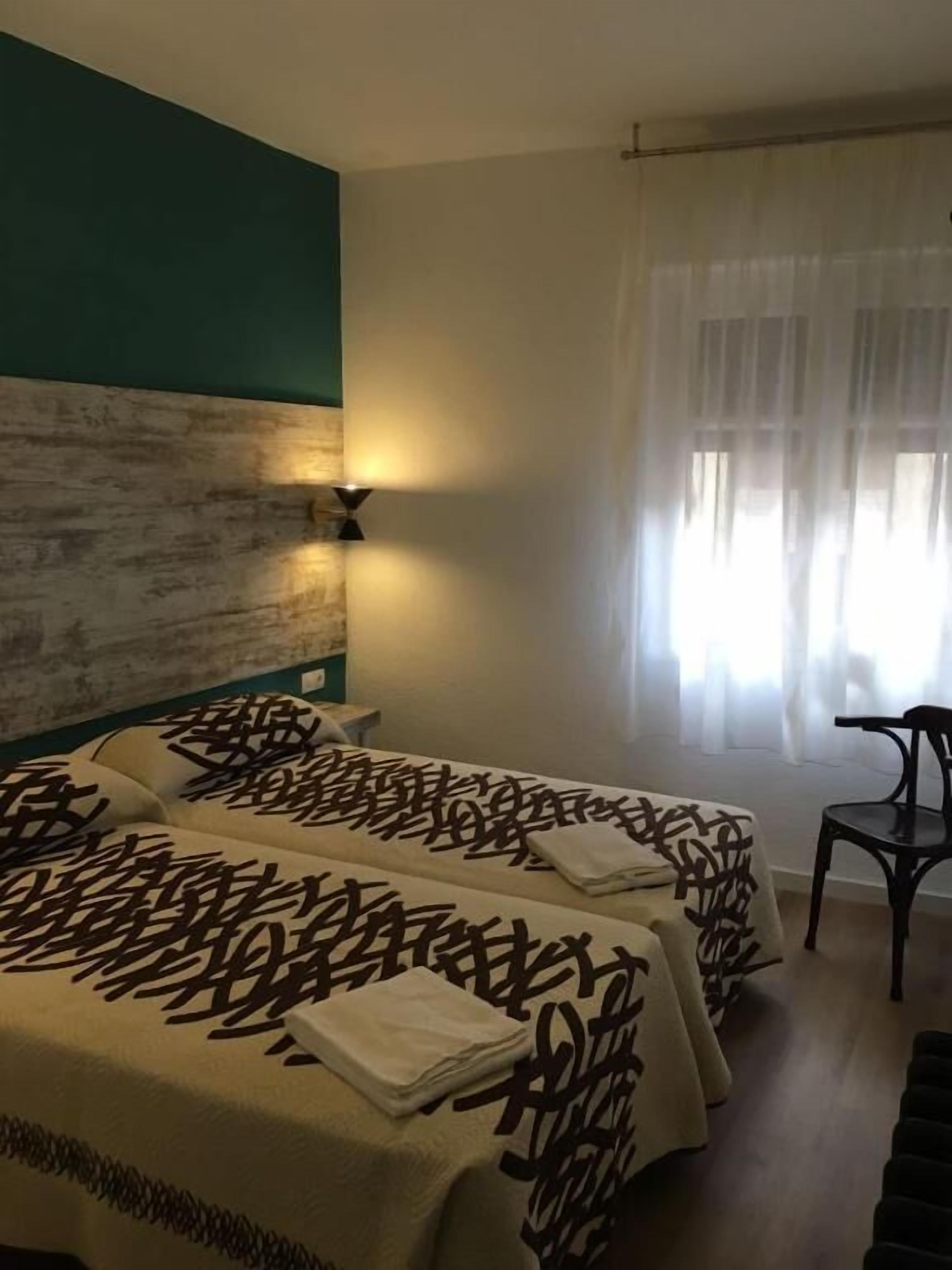 A hotel room with two beds, a wooden headboard, a wall-mounted lamp, a chair, and a window with sheer curtains.
