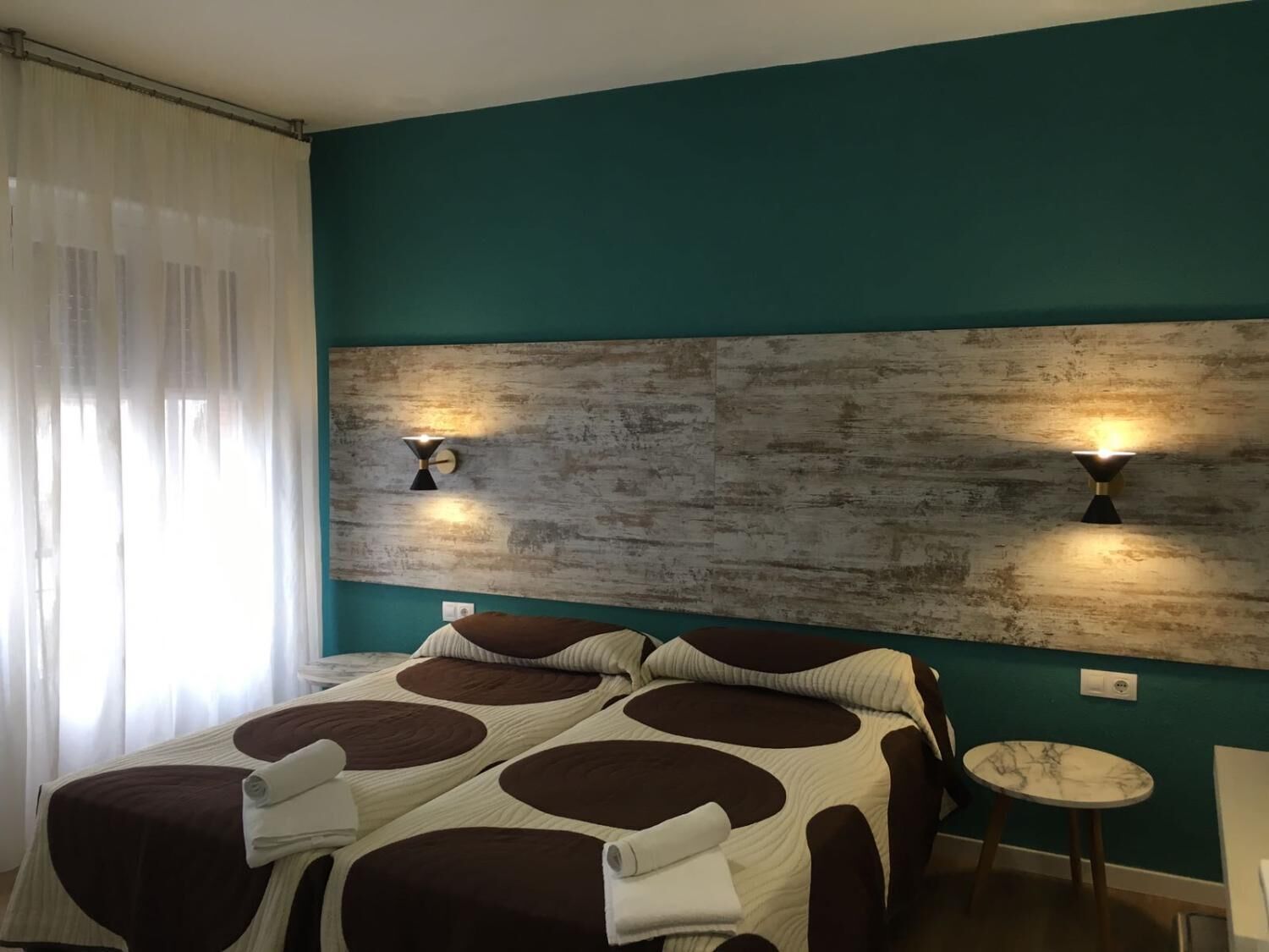Habitación de hotel con una cama, dos lámparas de pared, una mesita redonda y una cortina.