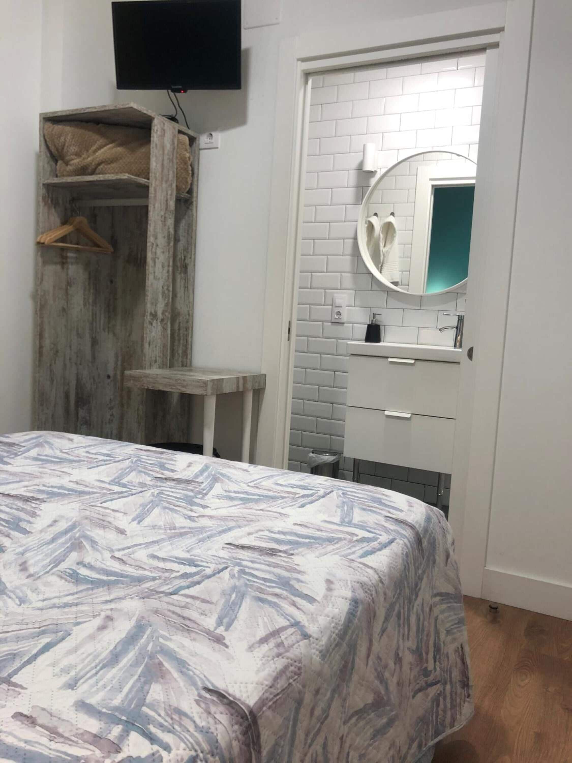 A bedroom with a bed, a wooden wardrobe, a small table, a mirror, and a television mounted on the wall.