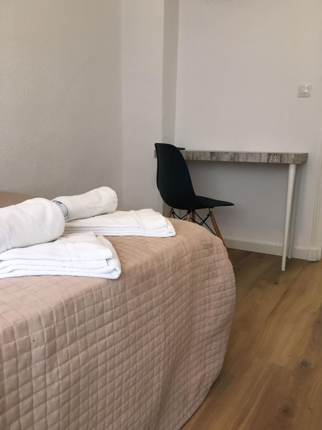 A neatly made bed with a pink quilted cover and folded white towels, a black chair, and a small wooden table against a plain wall.