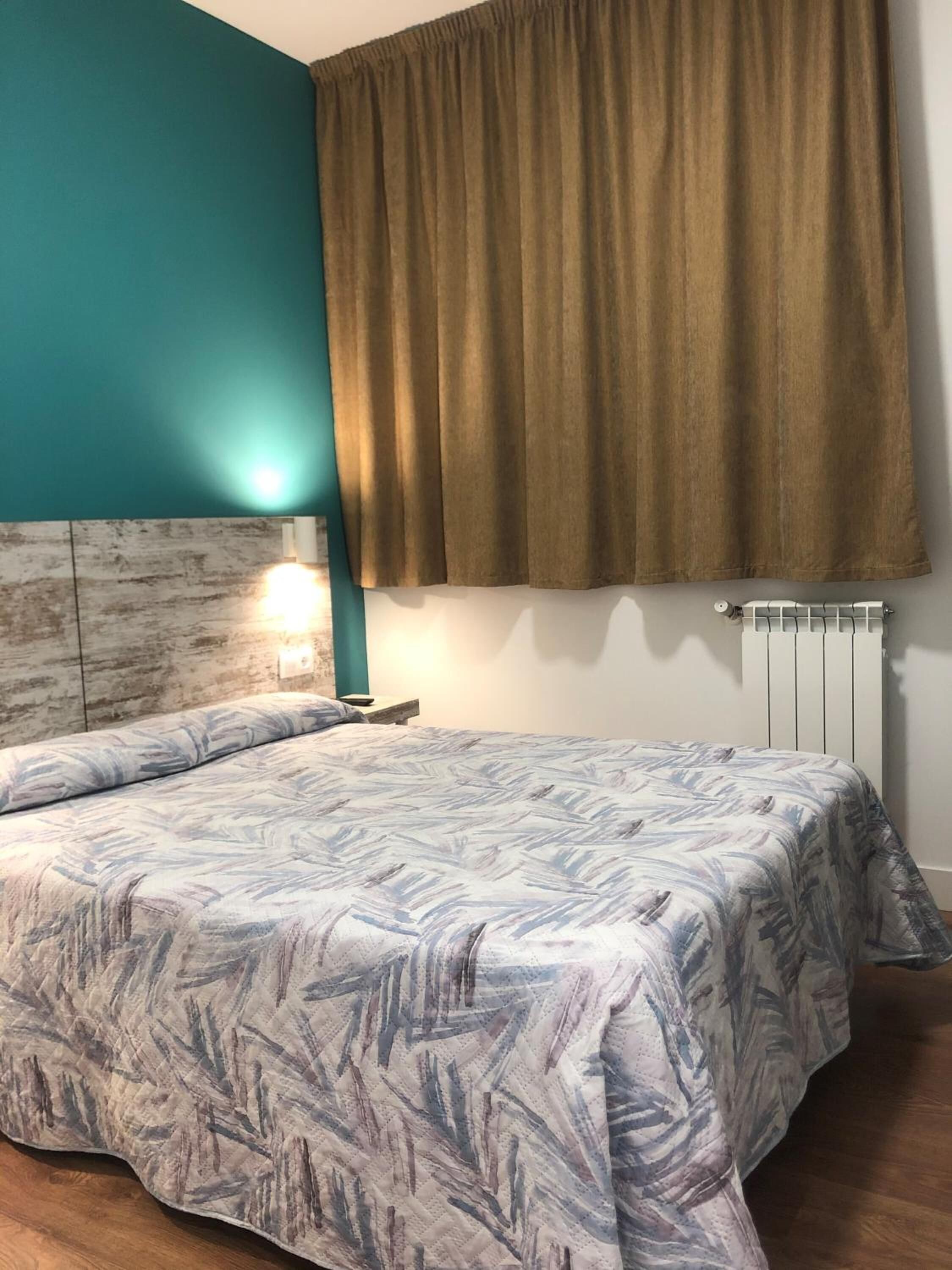 A bedroom with a bed, teal walls, a brown curtain, a white radiator, and a nightstand with a lamp.