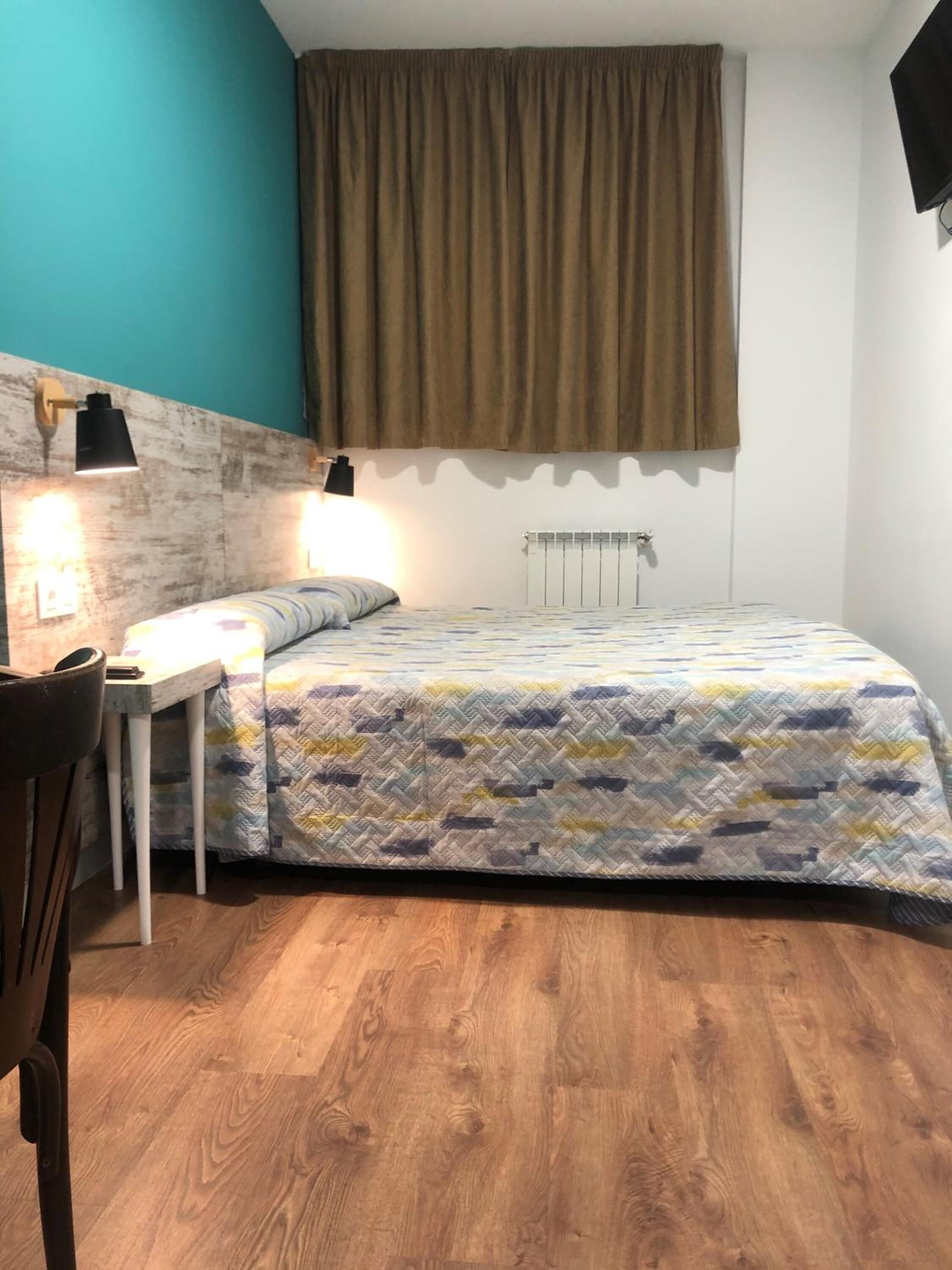 A bedroom with a bed, a desk, a chair, a window with curtains, and a radiator.