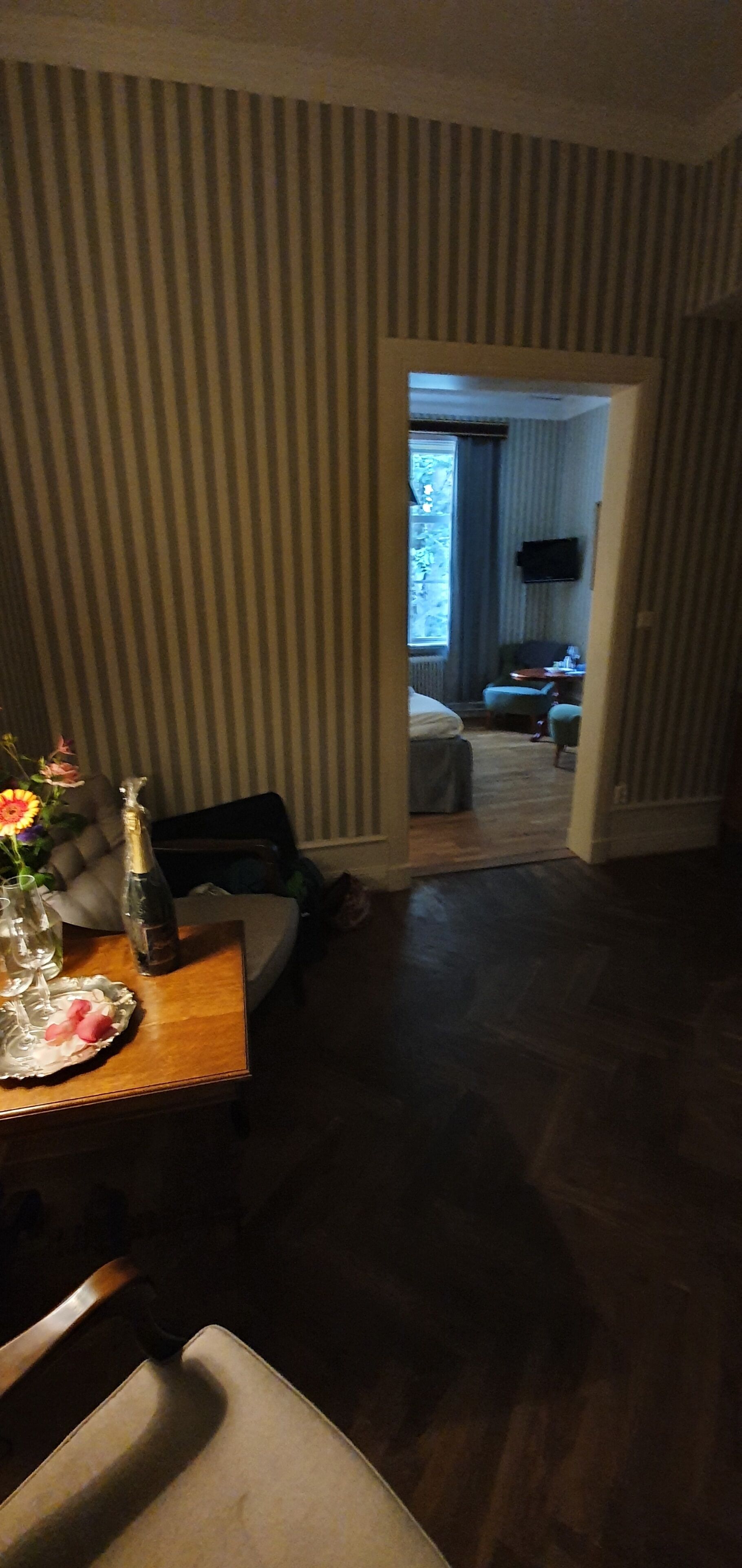 Ein Zimmer mit Streifenmuster-Tapete, ein Holzesstisch mit Blumenvase und Blick in ein weiteres Zimmer mit Bett und Fernseher.