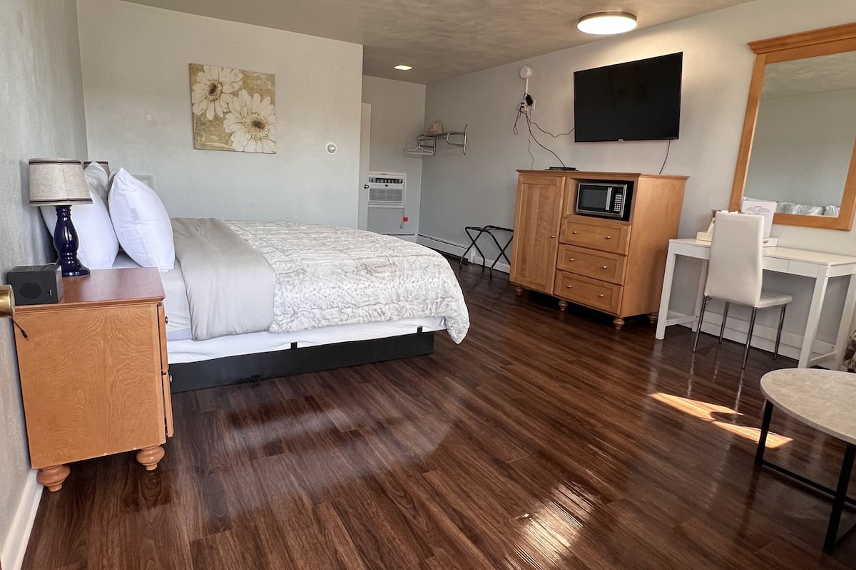 A bedroom with a bed, bedside table, lamp, dresser, microwave, TV, and a mirror.