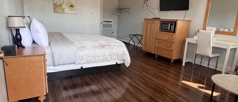 A bedroom with a bed, bedside table, lamp, dresser, microwave, TV, and a mirror.
