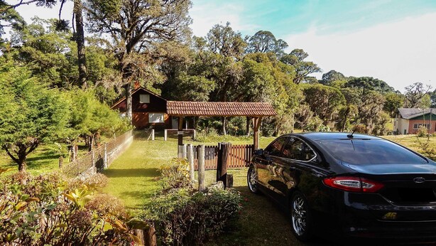 A black car parked in front of a wooden house with a tiled roof, surrounded by lush greenery and trees. - Chale Charm Gramado (Gramado)