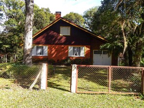 A rustic wooden house with a red roof, surrounded by lush greenery and a chain-link fence.