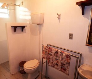 A small bathroom with a toilet, a sink, a towel rack with a floral towel, a wall-mounted light fixture, and a shelf above the toilet.