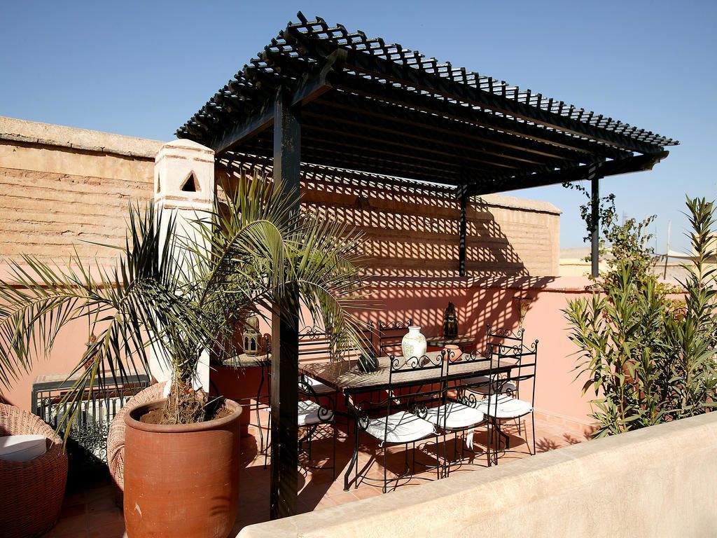 Een terras buiten met een pergola, een tafel met stoelen en potplanten.