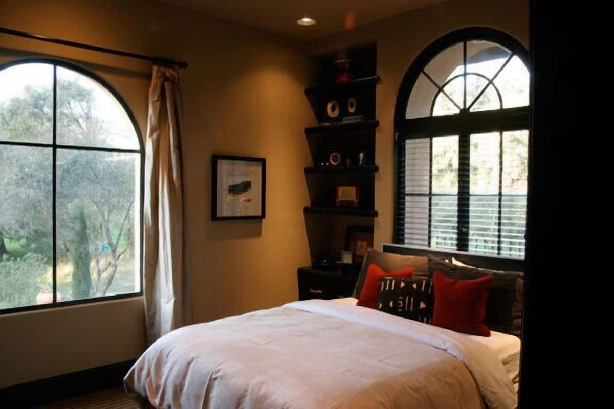 A bedroom with a large bed, a window with curtains, and a shelf with decorative items.