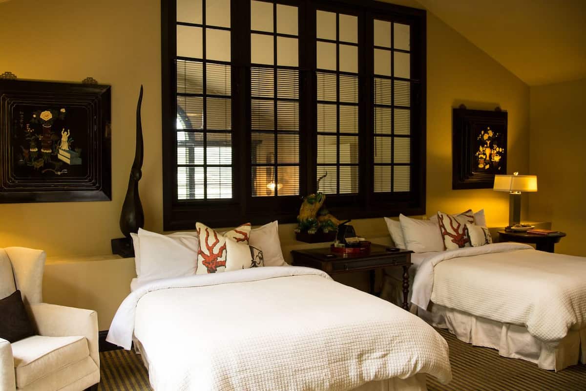 A hotel room with two beds, a chair, a large window, and artwork on the walls.