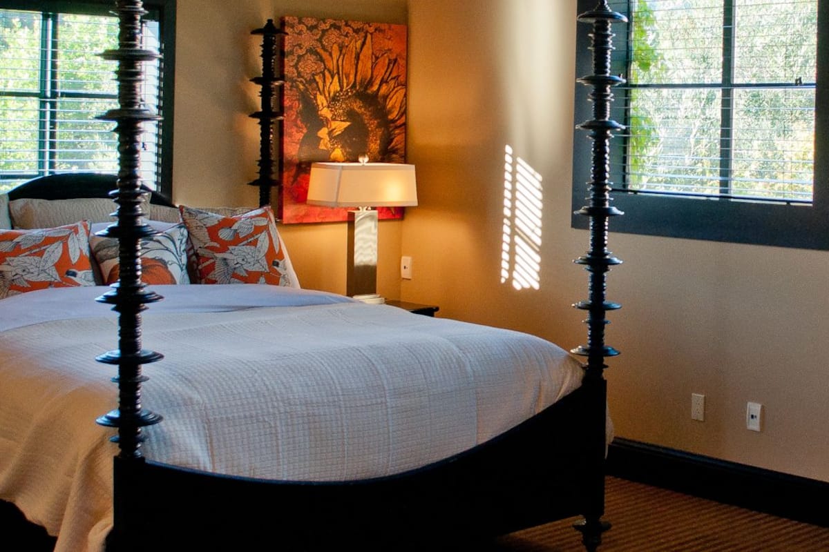 A four-poster bed with a white bedspread and orange and white patterned pillows. There is a nightstand with a lamp and a painting on the wall.