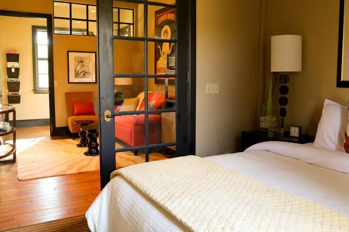 A hotel room with a bed, a nightstand, a lamp, a chair, and a door leading to another room.
