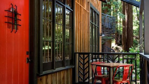 A wooden deck with a red door and black-framed windows, a small table, and two red chairs.