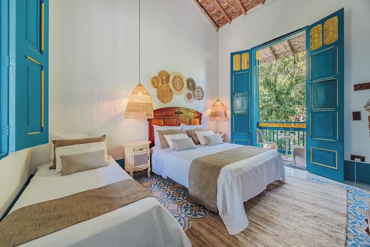 A room with two beds, a wooden headboard, woven wall decorations, and a view of greenery through open doors.