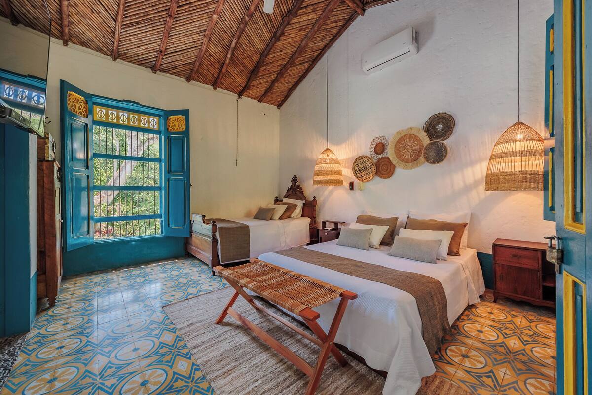 A room with two beds, a woven bench, a decorative wall, and a thatched ceiling.
