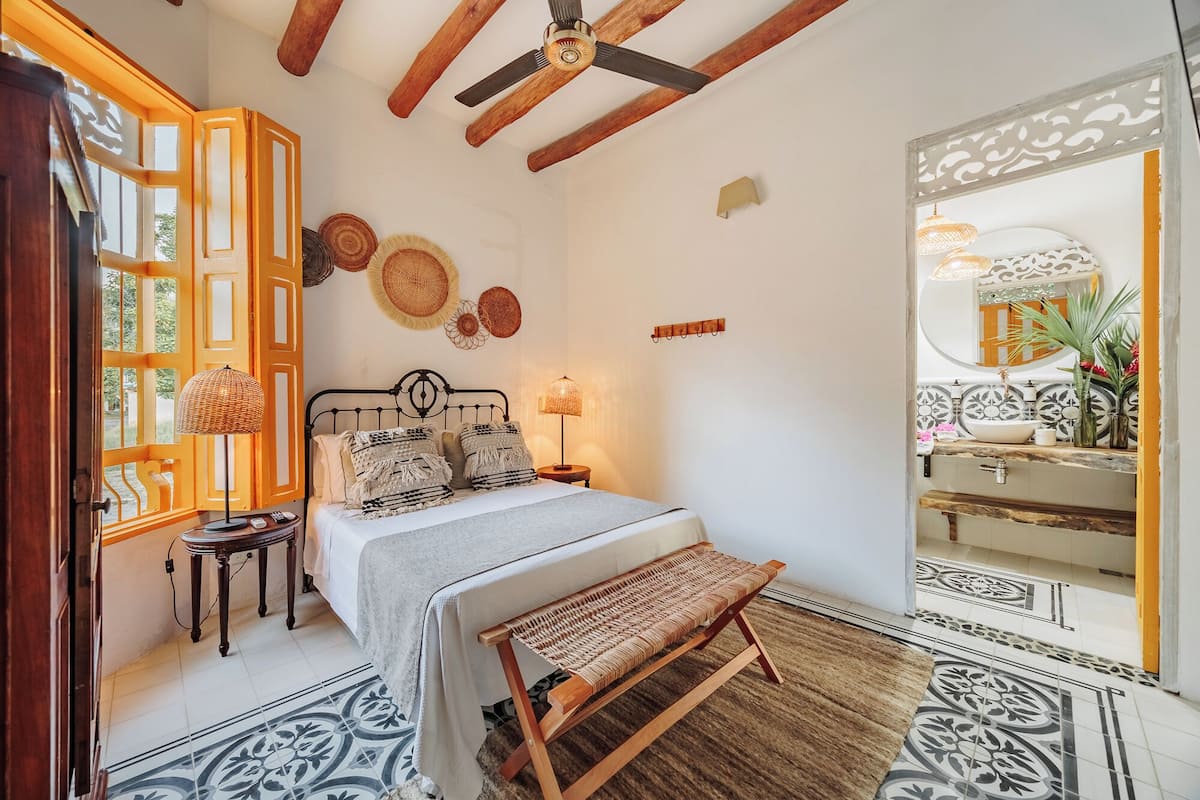 A bedroom with a bed, bedside lamps, a bench, a ceiling fan, and a bathroom with a mirror and sink.