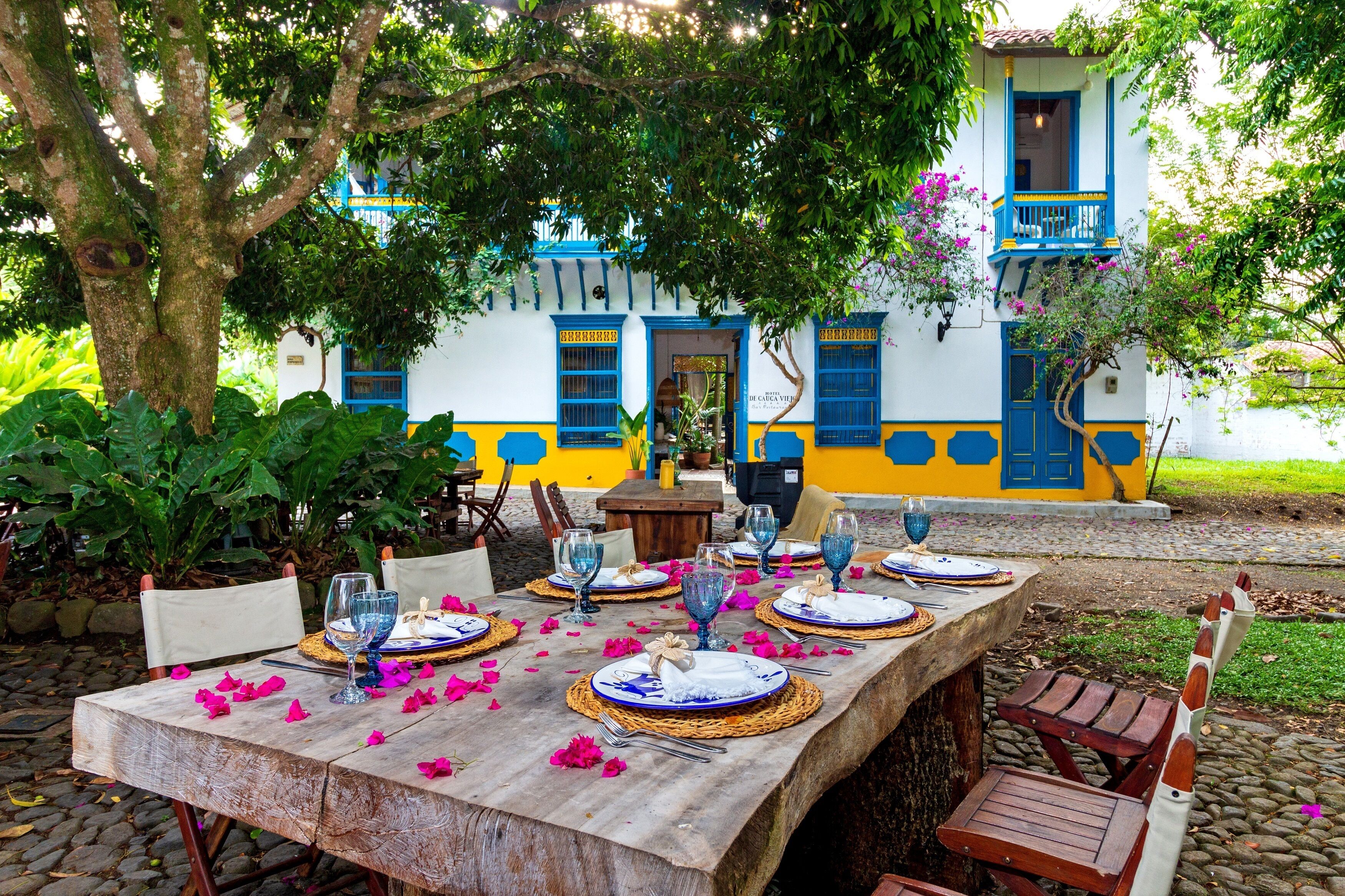 Un espace de restauration en plein air avec une table dressée pour quatre personnes, entourée de chaises, devant un bâtiment aux couleurs bleu et jaune.