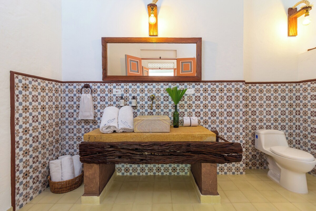 Een badkamer met een houten bankje, een wastafel met spiegel erboven, een toilet en een decoratieve wand met een ruitpatroon.