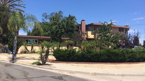 A two-story house with a brown roof and beige walls, surrounded by greenery and a clear blue sky.