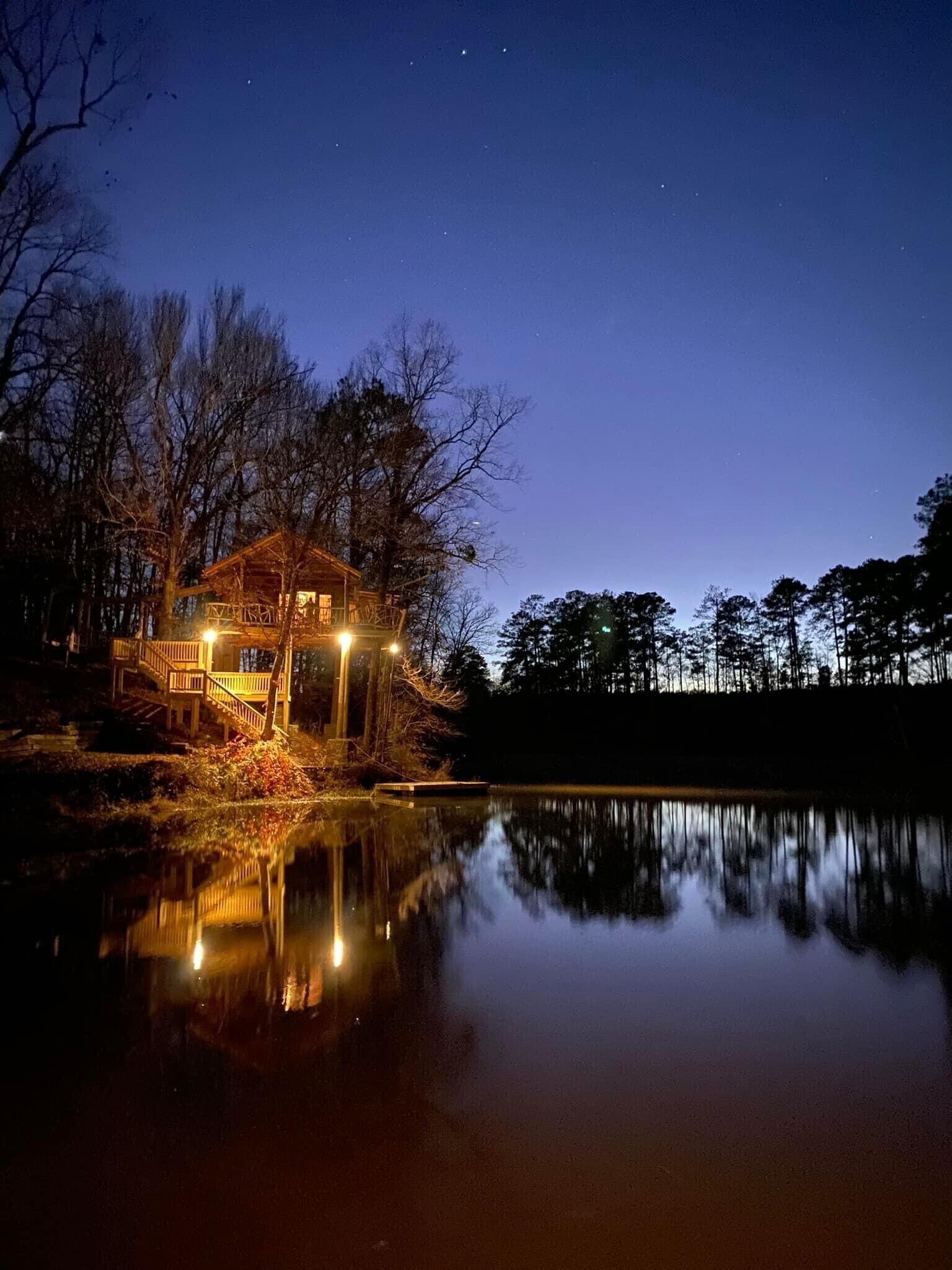 Una cabaña con terraza de madera y escalera, rodeada de árboles y un lago tranquilo que refleja la estructura.