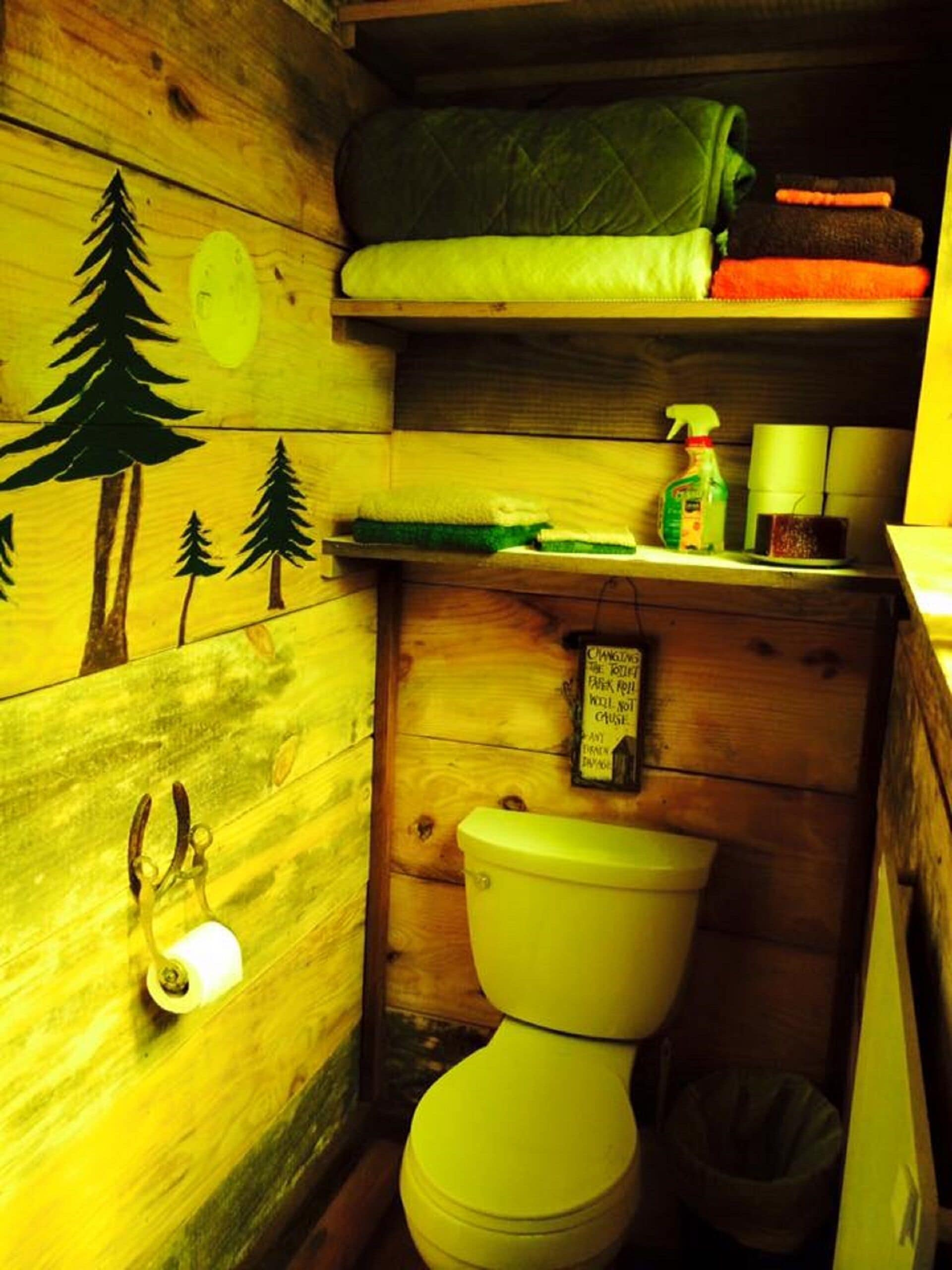 Een compacte houten badkamer met toilet, een klein plankje met toiletartikelen en een muurdecoratie van bomen.