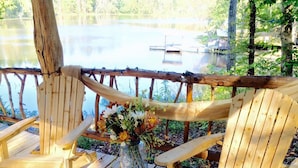 A wooden deck with chairs and a vase of flowers, overlooking a calm lake with a dock in the background. - 1 BEDROOM 1 BATH, TREE-CABIN BY A 20 ACRE PRIVATE LAKE (Jackson)