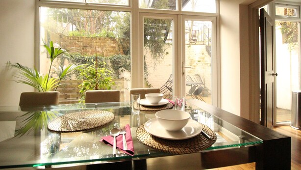 Una mesa de comedor dispuesta con platos, tazones y cubiertos, con vista a un jardín a través de amplios ventanales.