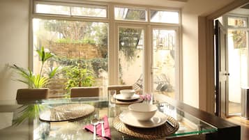 A dining table set with plates, bowls, and cutlery, with a view of a garden through large windows.