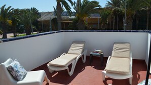 Een zonnig terras buiten met twee lounge fauteuils en een kleine tafel, omringd door palmbomen en gebouwen op de achtergrond.