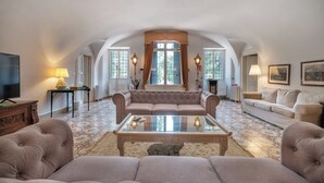 Ein großzügiger Wohnraum mit hoher Decke, einem großen Fenster mit Vorhängen, einem Sofa, einem Couchtisch und einem Fernseher.