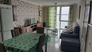 A compact living space with a dining area, a sofa, a television, and a view of the sea through large windows.