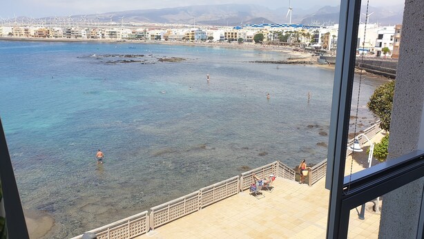 A coastal view from a balcony with clear water, a beach, and wind turbines in the background.