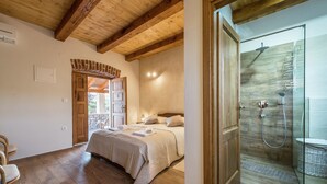 Une chambre avec un plafond en bois, un lit recouvert d’une couverture beige, une porte en bois et un espace douche doté d’une porte en verre.