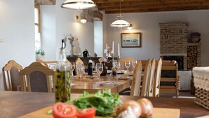 Une salle à manger avec une table en bois dressée pour un repas, entourée de chaises. On aperçoit une cheminée au fond et un banc en bois sur la droite.