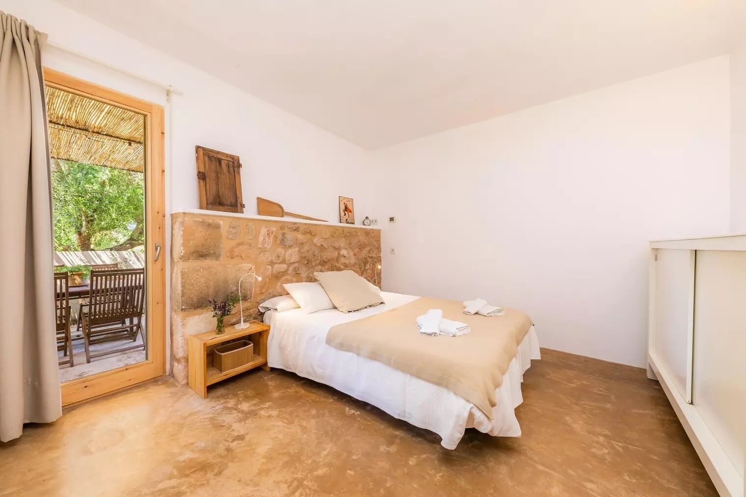 A bedroom with a stone headboard, a bed with white linens, a bedside table with a lamp, and a view of an outdoor patio.