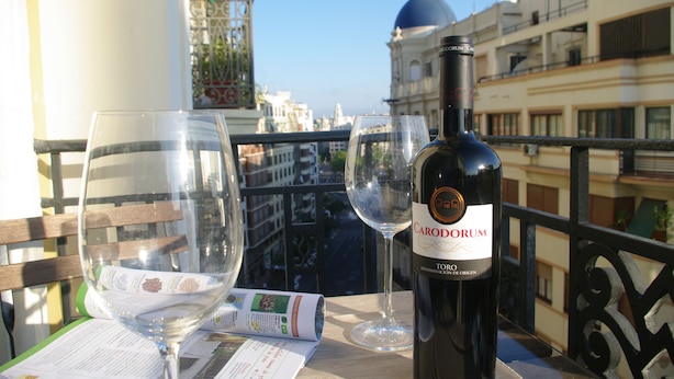 Une bouteille de vin Caro Dorum, deux verres à vin vides et un menu sur une table de balcon, avec une vue sur la ville en arrière-plan.