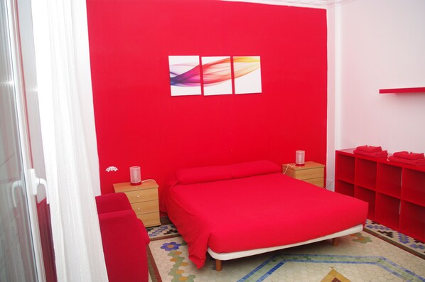 A bedroom with a red wall, a bed with a red bedspread, a nightstand with a lamp, a dresser, and a red shelving unit.
