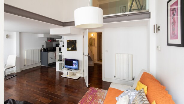A modern living room with a flat-screen TV, a sofa with orange cushions, and a white pendant light. - A Stylish 2 Bed, 2 Bath London Apartment (London)