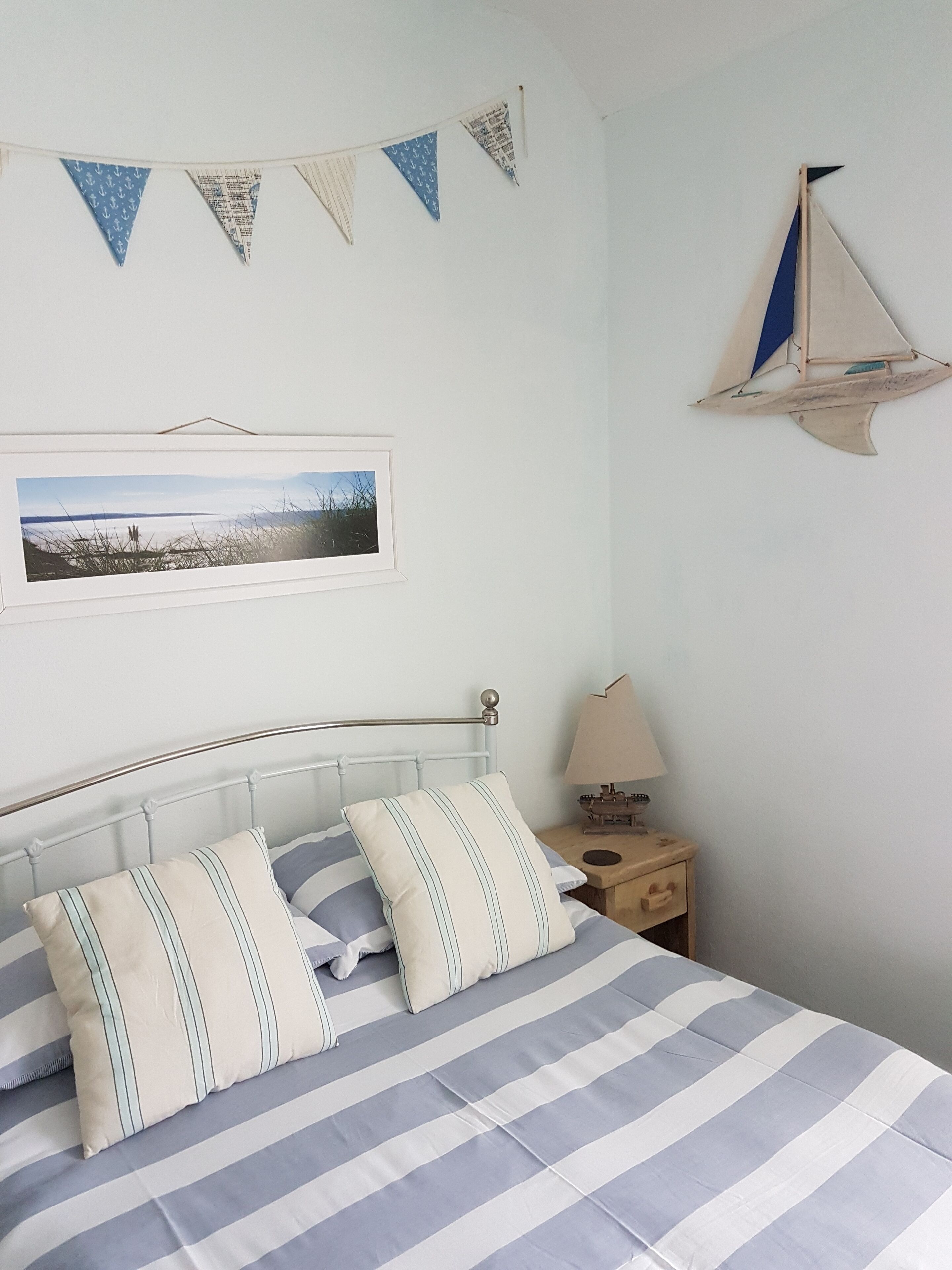 Een netjes opgemaakt bed met gestreept beddengoed, een ingelijste foto aan de muur en een klein houten nachtkastje met een lamp.